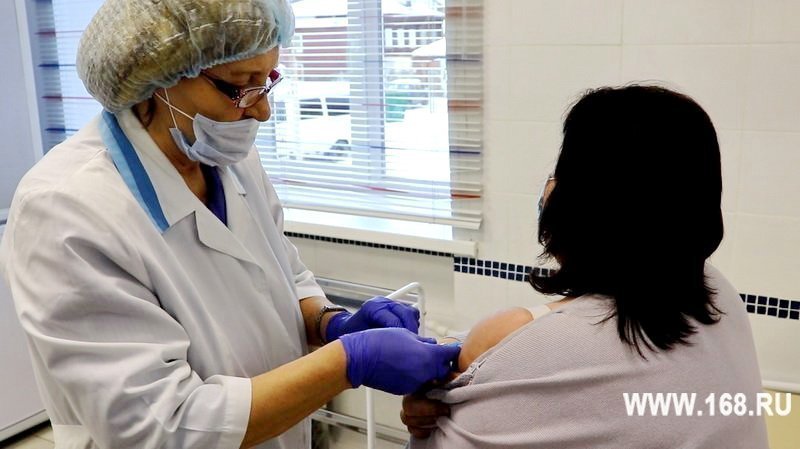 медработник в маске и шапочке делает укол в плечо сидящей пациентке в медицинском кабинете, водяной знак WWW.168.RU