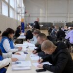 Внутренний вид спортивного зала: несколько рядов столов, люди заполняют бланки; волонтёры/организаторы в синих жилетах сидят за столами.