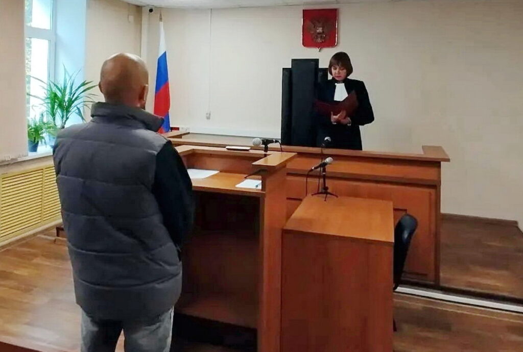 Мужчина стоит перед судьёй, читающей документ у стола в зале суда с российским флагом и гербом.