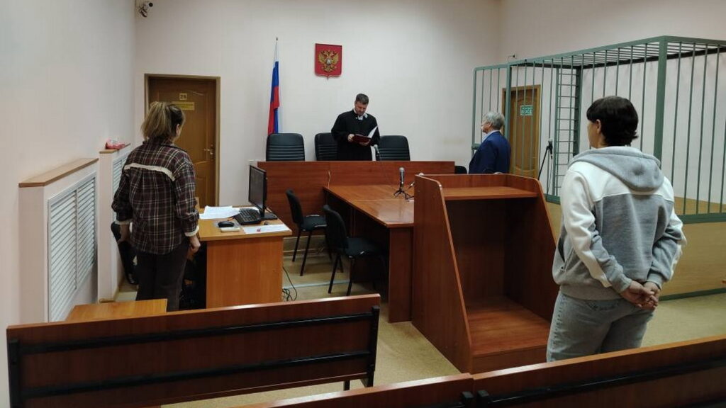 Зал суда: мужчина в мантии читает документ, перед ним стоят трое участников заседания, справа видна металлическая клетка.