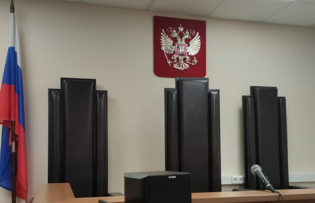 Зал суда с тремя высокими креслами, российским флагом, гербом и микрофоном
