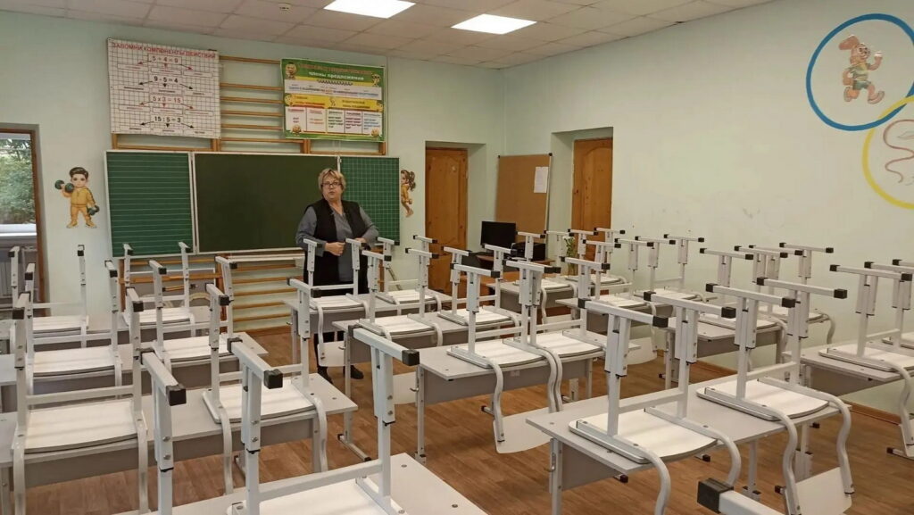 Женщина стоит в пустом учебном классе, где стулья перевёрнуты и поставлены на парты