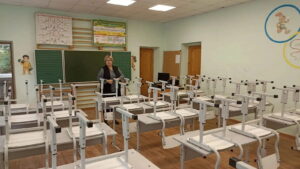 Женщина стоит в пустом учебном классе, где стулья перевёрнуты и поставлены на парты