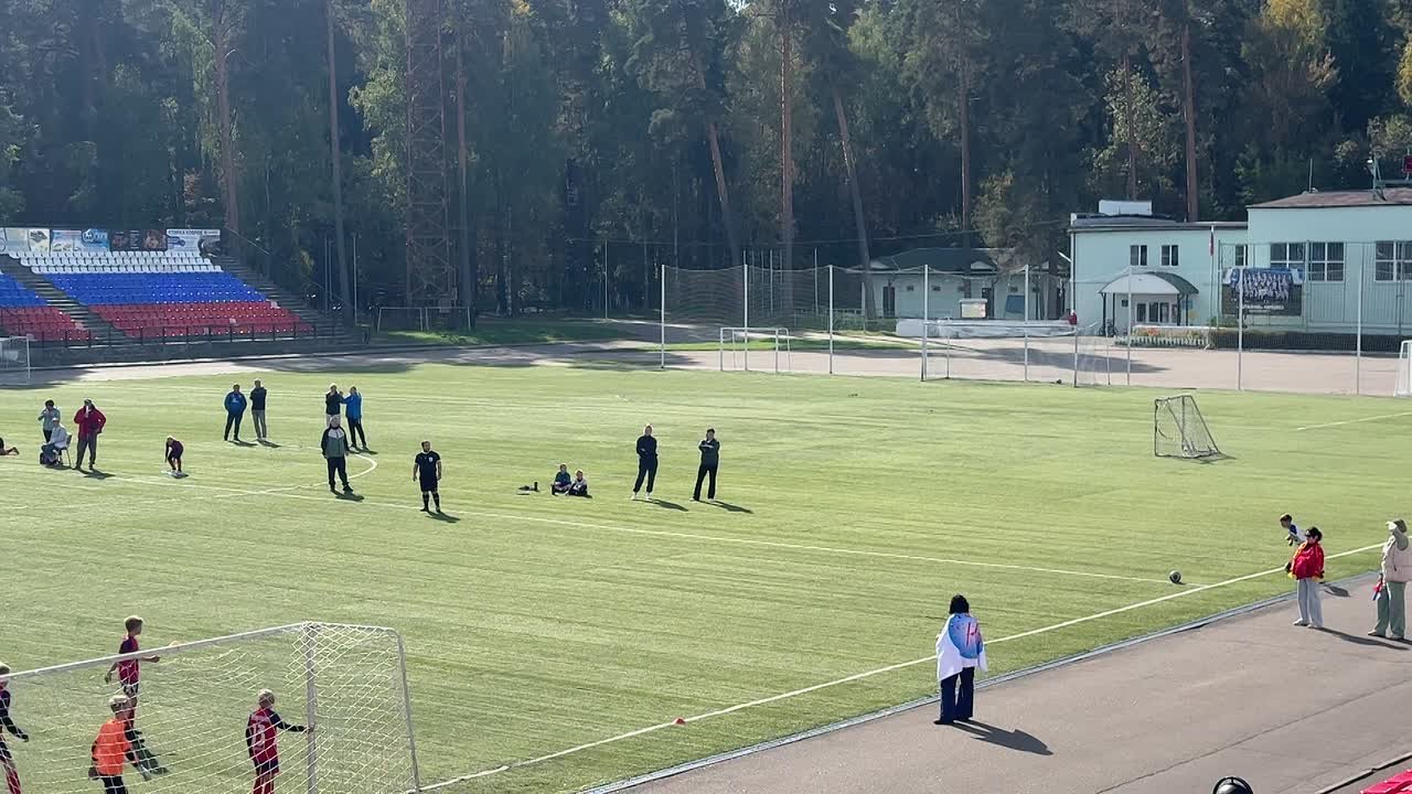 Футбольное поле во время матча: игроки на газоне, трибуна слева, мяч у угловой зоны и ворота в кадре.