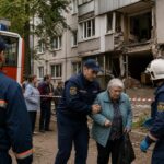 Спасатели помогают пожилой женщине на фоне повреждённого многоквартирного дома и пожарной машины