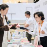 Столы с книгами в торговом атриуме; продавец/волонтёр с бейджем обслуживает посетительницу, на заднем плане ещё один посетитель и стеллажи.