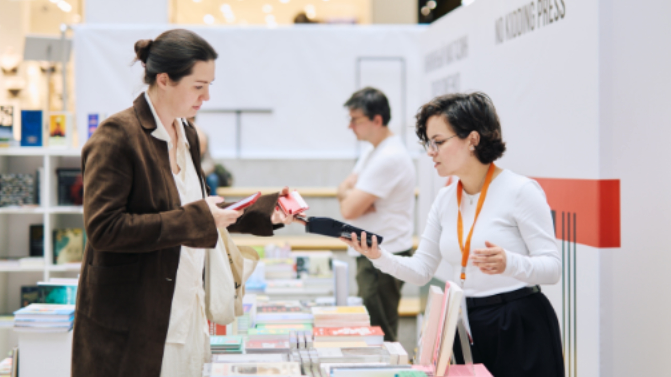 Столы с книгами в торговом атриуме; продавец/волонтёр с бейджем обслуживает посетительницу, на заднем плане ещё один посетитель и стеллажи.