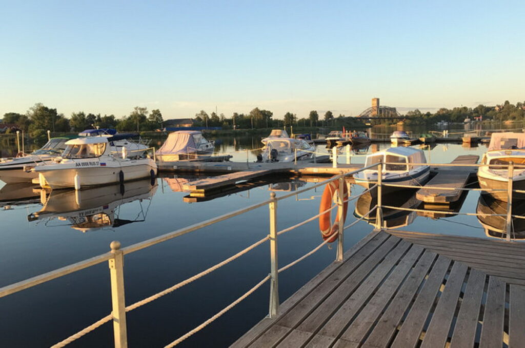 Моторные лодки и катера у плавучих причалов на спокойной воде, на переднем плане деревянный настил с ограждением и спасательным кругом