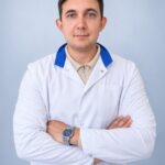 портрет молодого врача в белом халате, стоит с сомкнутыми руками на светлом однотонном фоне