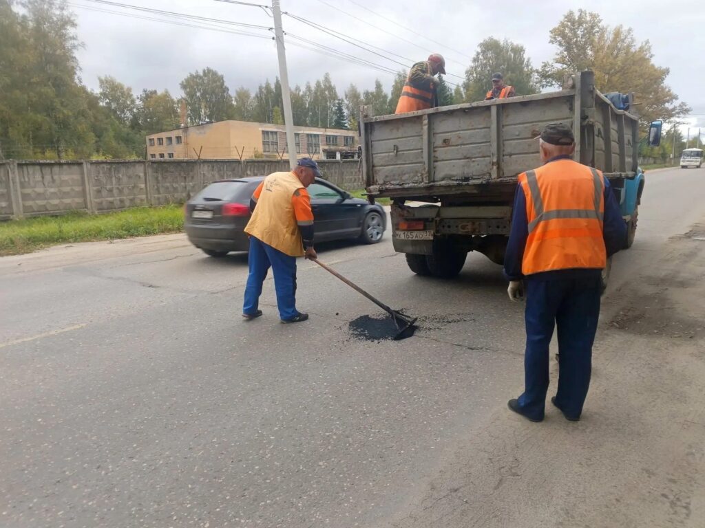 Дорожные рабочие возле грузовика; один из них разравнивает асфальт в выбоине