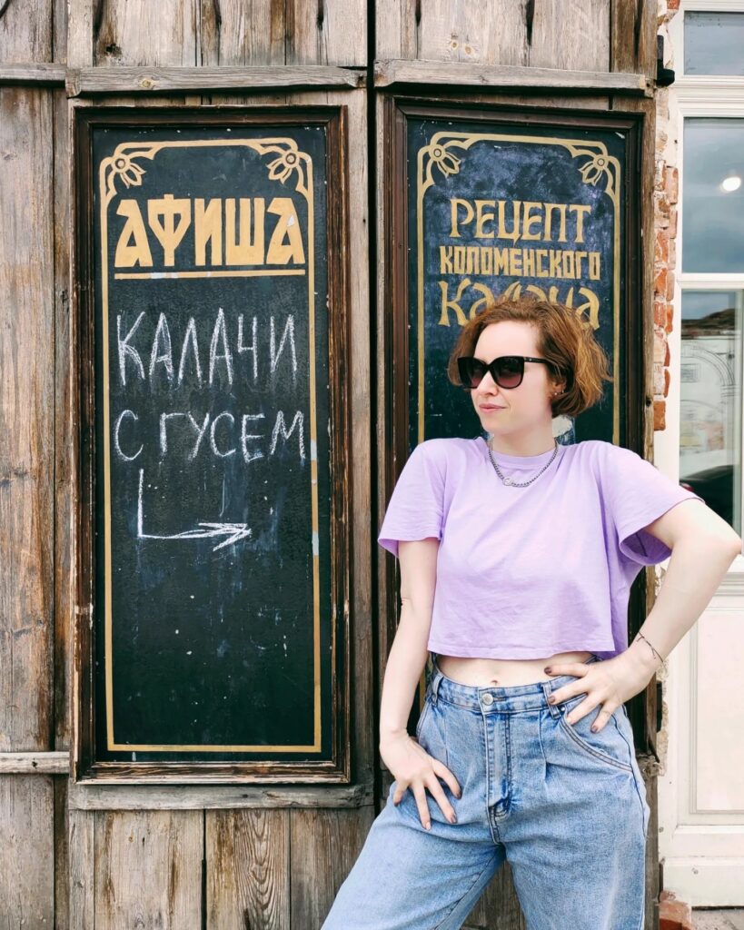 Женщина в тёмных очках позирует у деревянной стены с двумя чёрными досками и надписями мелом