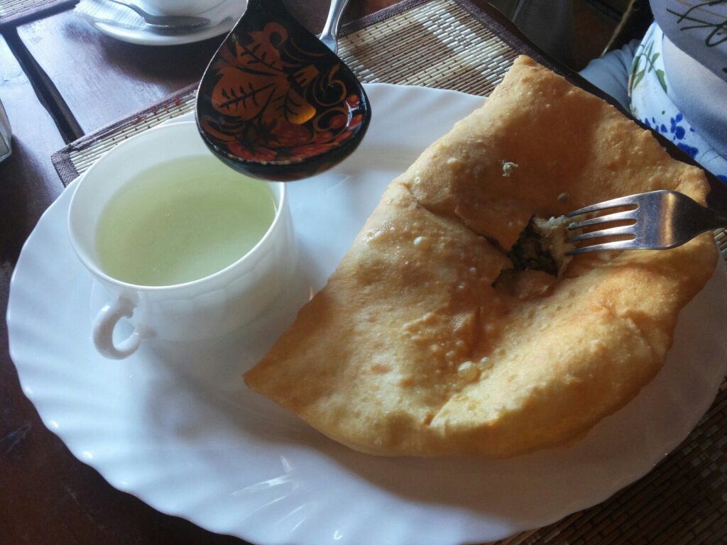 Большой жареный пирог с мясной начинкой и вилка лежат на тарелке рядом с чашкой светлого бульона