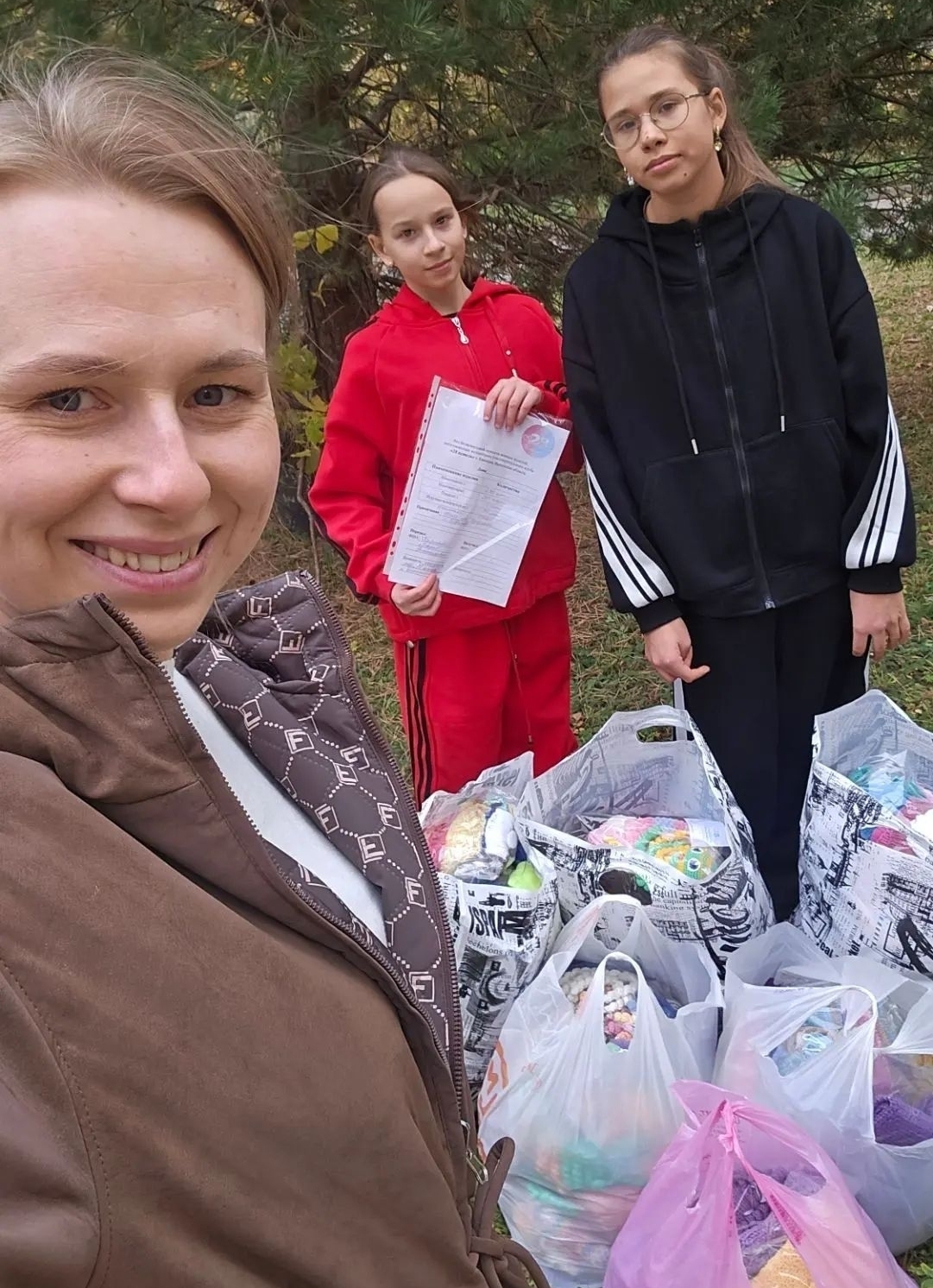 три женщины/девушки на открытом воздухе рядом с множеством пакетов и сумок, в которых видны вязаные вещи