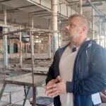 Мужчина с микрофоном стоит в пустом цехе с облупившимися колоннами и остатками оборудования