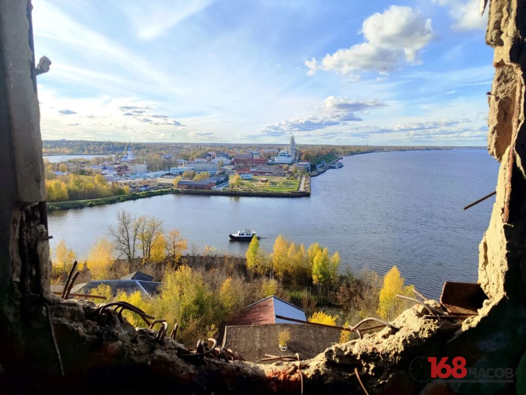 Вид через разрушенный проём на широкую реку, осенний город и небольшой катер