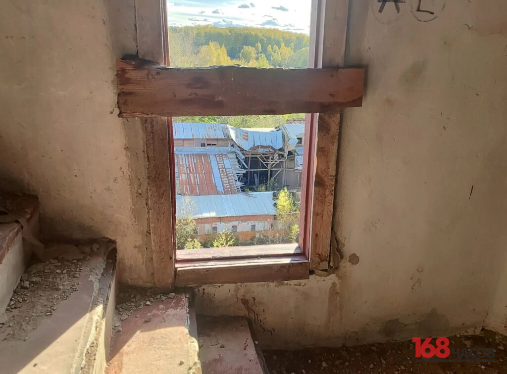 Заколоченное доской окно в обветшавшем помещении, за ним повреждённые крыши промышленных корпусов