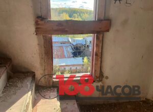 Заколоченное доской окно в обветшавшем помещении, за ним повреждённые крыши промышленных корпусов