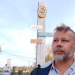 Седой мужчина на фоне реки и высокой стелы с гербом и надписью «Слава орденоносному городу»