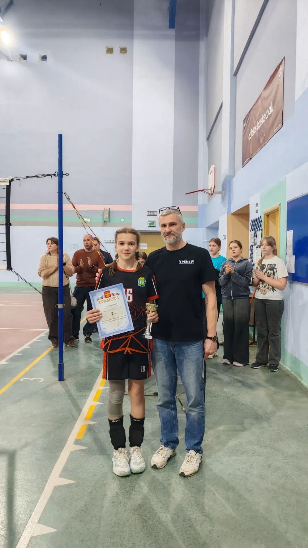 Девочка в волейбольной форме держит грамоту и кубок рядом с мужчиной в футболке с надписью «ТРЕНЕР» в спортивном зале; на заднем плане сетка и аплодирующие участники.