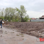 пустой грязный участок земли с колеями и лужами, рядом металлический забор и малоэтажные дома, слева стоит человек