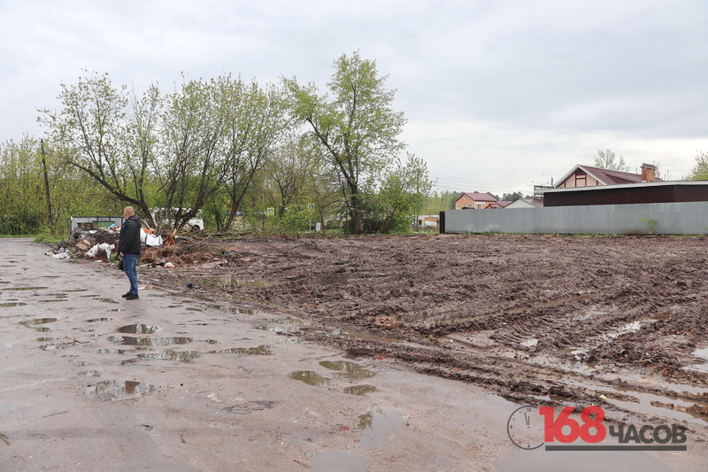 пустой грязный участок земли с колеями и лужами, рядом металлический забор и малоэтажные дома, слева стоит человек