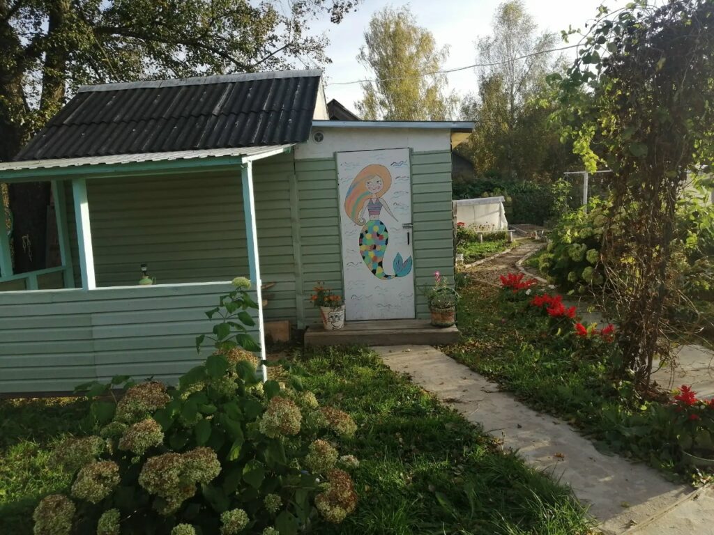 Небольшая зелёная постройка в саду, на белой двери которой нарисована русалка с разноцветным хвостом