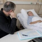 Мужчина сидит у кровати пациента в палате стационара; на столике перед ним папка с бланком и ручка.