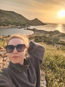 Женщина в крупных тёмных очках делает селфи на фоне скалистой бухты и закатного солнца