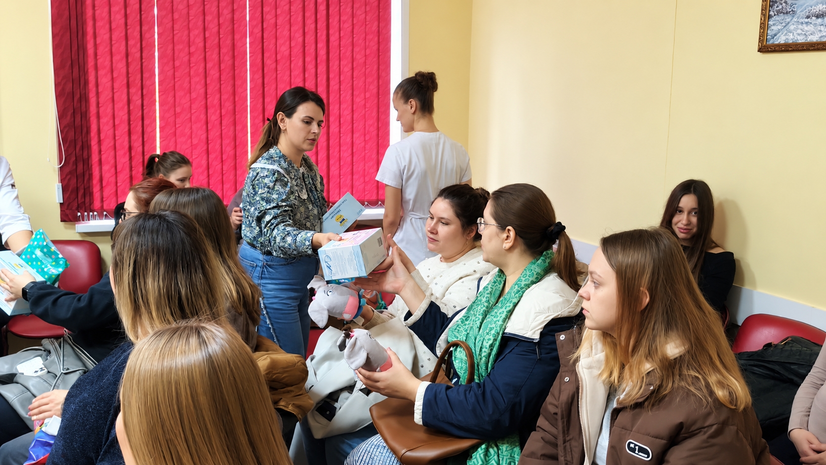 Группа женщин в актовом зале (клиническое помещение) получает подарки; женщина раздаёт коробки и мягкие игрушки, на заднем плане медработник в белом халате.