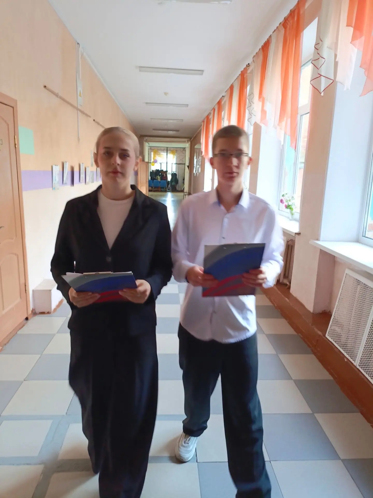 Двое подростков идут по школьному коридору, держат папки/планшеты в руках