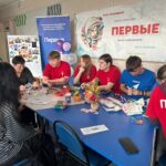 Студенты и школьники за столом с поделками и материалами на мастер-классе; на заднем плане видны вертикальный синий баннер и большой баннер с надписью «Первые».