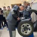 Школьники и студенты в рабочей форме вокруг автослесарного станка с колесом — демонстрация в мастерской
