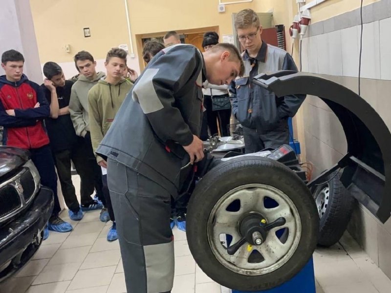 Школьники и студенты в рабочей форме вокруг автослесарного станка с колесом — демонстрация в мастерской