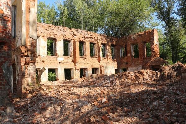 Разрушенный кирпичный двухэтажный дом с обрушенными стенами и завалом кирпича на переднем плане, вокруг — деревья.