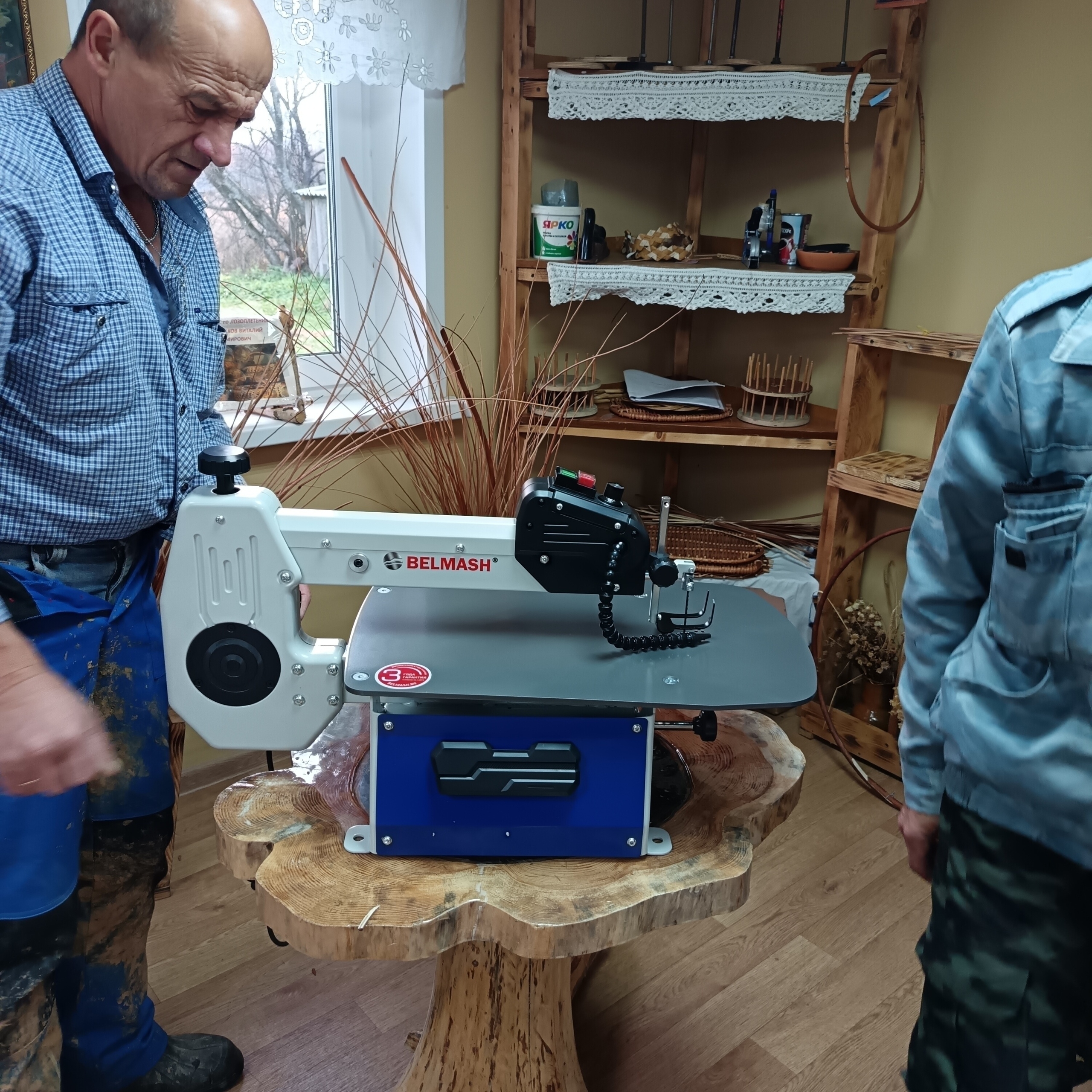 Мужчины рядом с новым станком марки BELMASH в ремесленной мастерской; на полках — плетёные поделки и мастерские принадлежности.