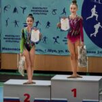 Две юные гимнастки на пьедестале с грамотами и медалями на фоне баннера «ФАКУЛЬТЕТ ФИЗИЧЕСКОЙ КУЛЬТУРЫ»