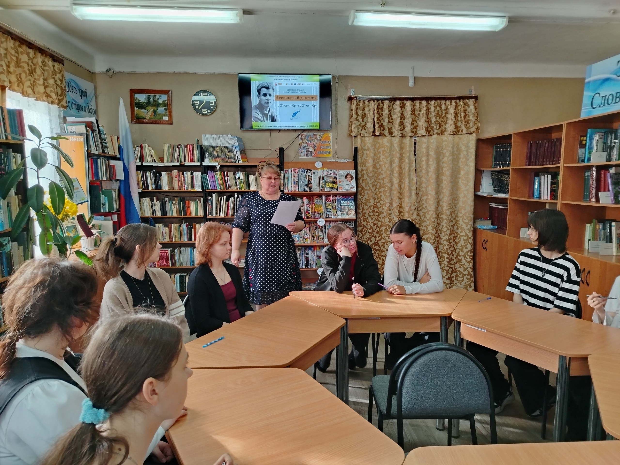 Группа школьников за столами в библиотеке; преподаватель стоит с листом на фоне книжных полок и экрана с афишей «Есенинский диктант».