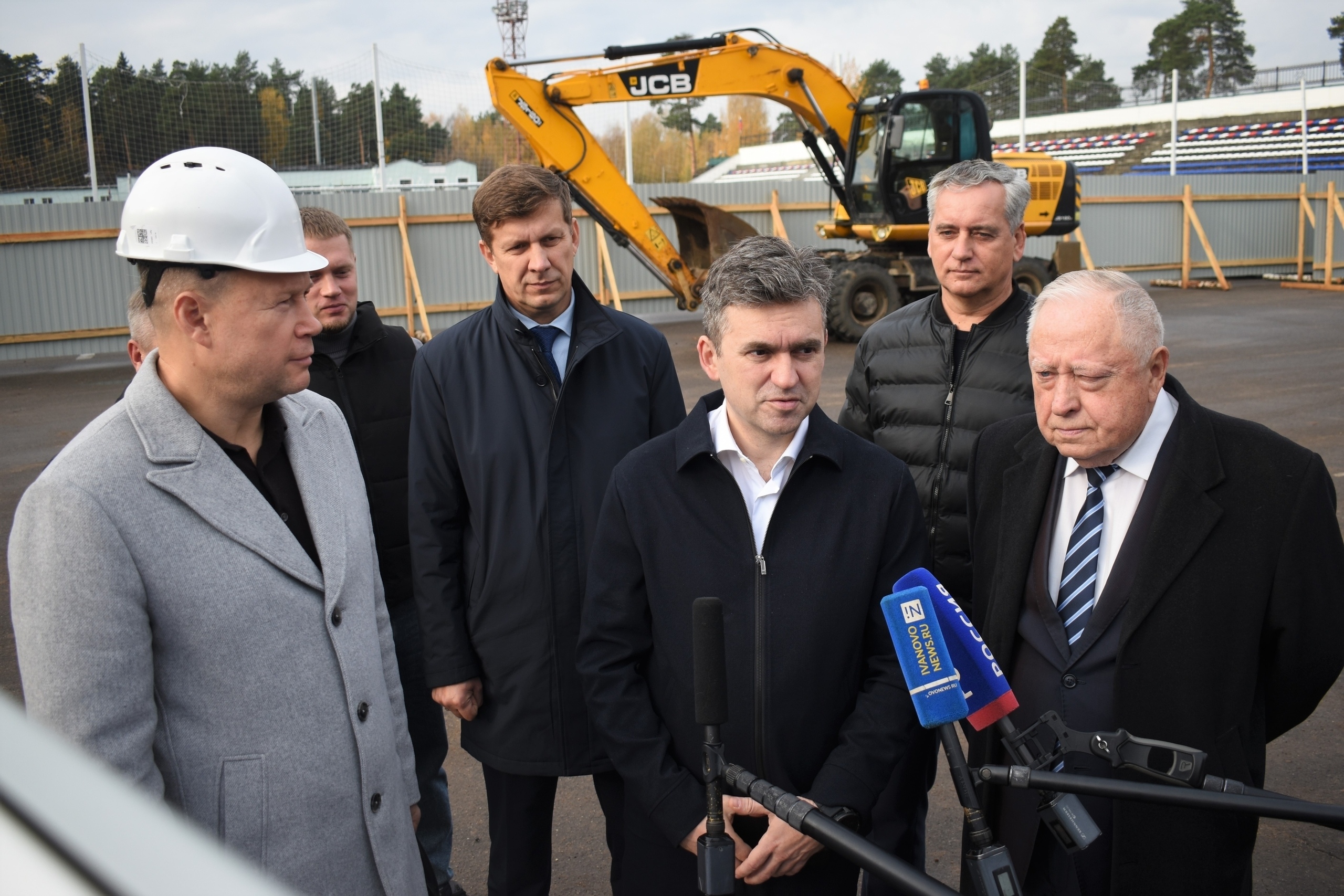 Несколько мужчин у строительной площадки: экскаватор JCB, временное ограждение стройплощадки и трибуны стадиона на заднем плане; микрофоны на переднем плане.