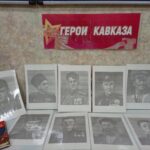 Плакат с надписью «Герои Кавказа» над рядами чёрно‑белых портретов и книгой на столе