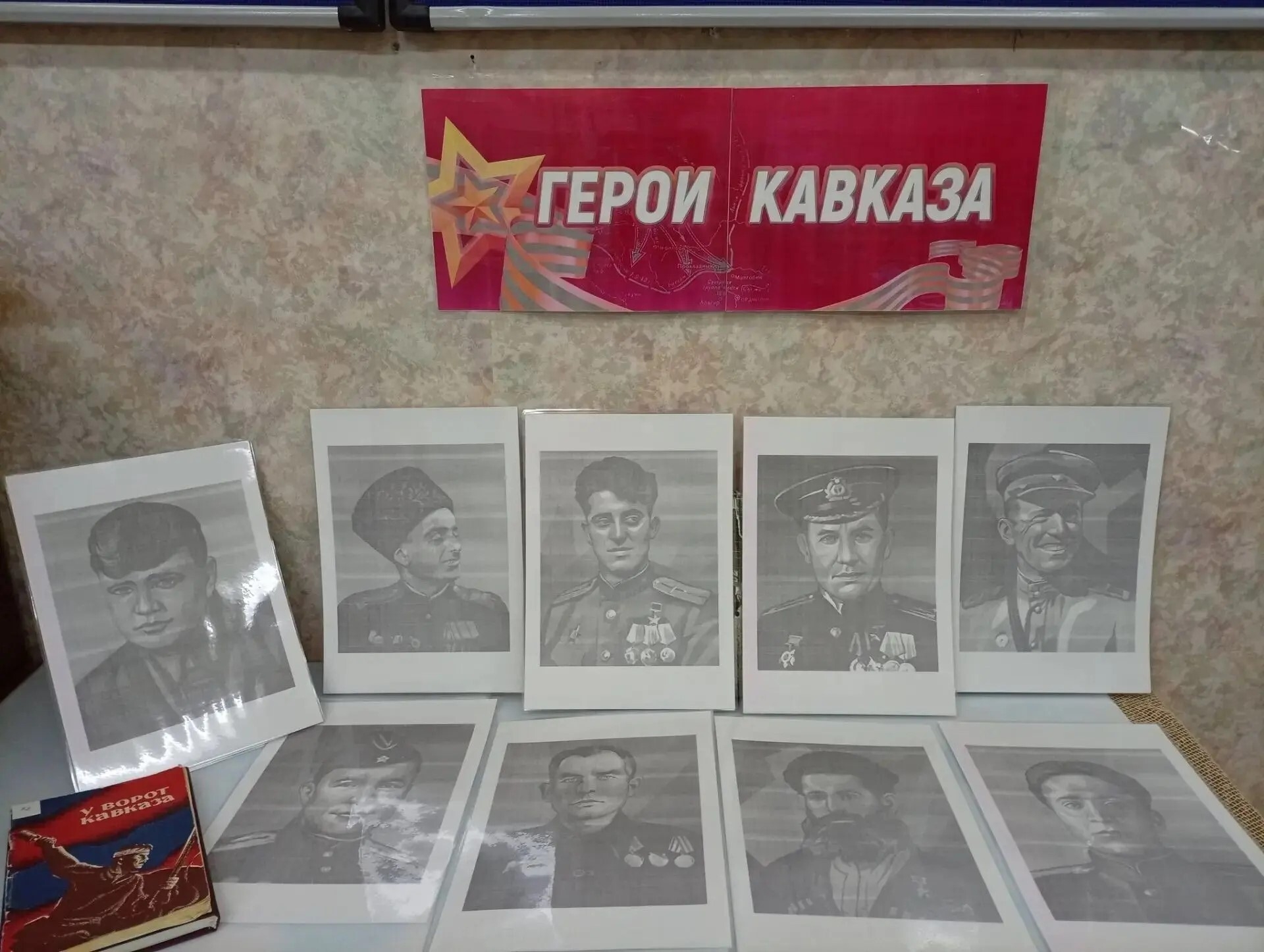 Плакат с надписью «Герои Кавказа» над рядами чёрно‑белых портретов и книгой на столе