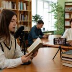 В читальном зале библиотеки девушка читает вслух в микрофон; на столе смартфон на штативе записывает, вокруг книжные полки и другие читатели.