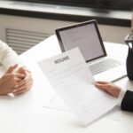 Собеседование: на столе лист с заголовком «RESUME», ноутбук и руки рекрутера и соискателя