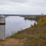 Берег реки с бетонным пограничным столбом, на котором нанесены красная и зелёная полосы; на противоположном берегу видны сельские дома и церковь, осенний пейзаж.