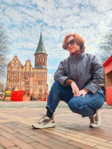 Женщина в тёмных очках сидит на корточках на площади перед кирпичным зданием с часовой башней
