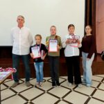 Церемония награждения: четыре ребёнка (девочки) с дипломами, медалями, кубком и подарочными наборами на сцене вместе с двумя взрослыми.