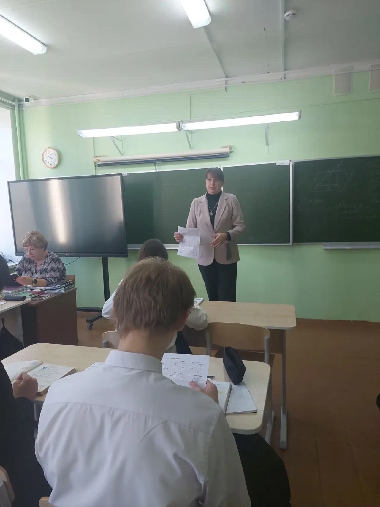 Взрослая женщина стоит у школьной доски и держит раздаточные листы; перед ней – несколько школьников за партами; в классе видны интерактивный экран и мелованная доска.