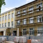 Фасад школы в строительных лесах при капитальном ремонте, на переднем плане стройматериалы и рабочие