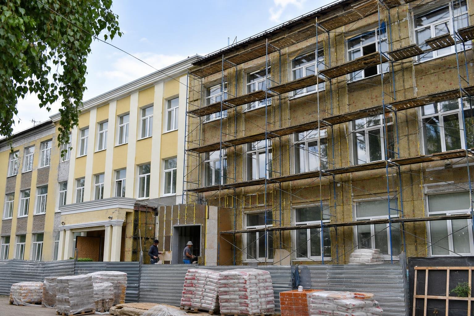 Фасад школы в строительных лесах при капитальном ремонте, на переднем плане стройматериалы и рабочие