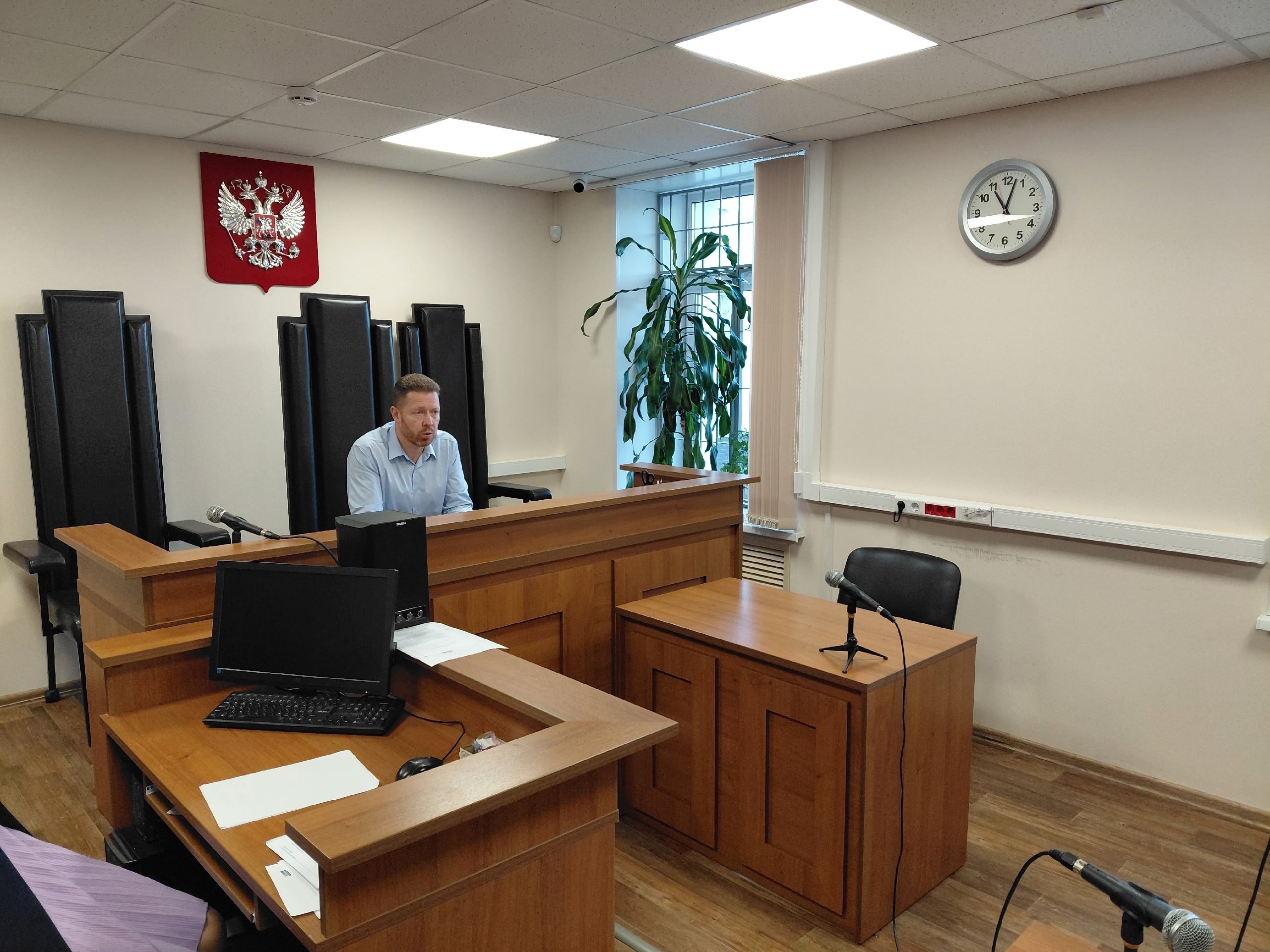 интерьер зала судебного заседания: судейская кафедра с гербом на стене, деревянные столы и микрофоны; мужчина сидит за столом