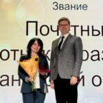 Женщина с букетом и футляром награды стоит на сцене рядом с мужчиной; на фоне проекционный экран с крупными словами «Звание», «Почетный» и частью слова «работник/Иван…»
